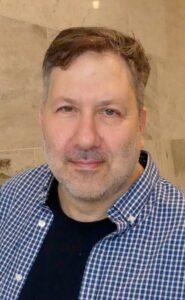 Bilden är en ansiktsbild på en man klädd i svart t-shirt under en blåvitrutig skjorta.